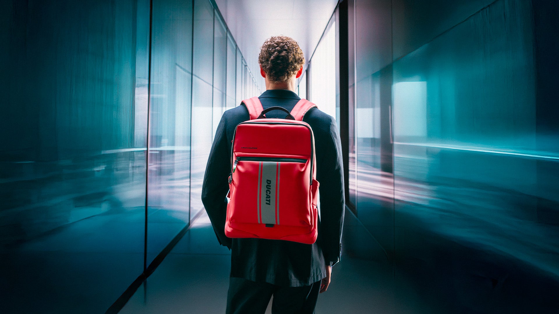 Person seen from behind walking through a futuristic glass corridor, wearing a bright red Ducati-branded backpack.